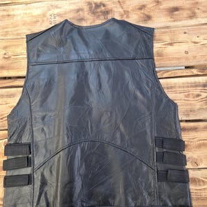 May include: A black leather vest with a patchwork design. The vest has a V-neck and features three black straps on each side. The vest is displayed on a wooden surface.