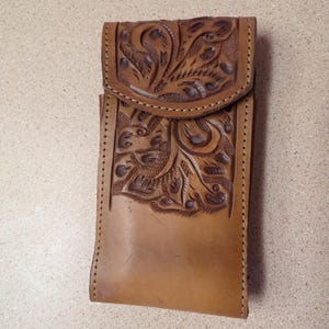 May include: A tan leather case with a flap closure. The case features detailed floral and leaf carvings on the front and flap. The edges are stitched with a contrasting thread, and the leather has a natural, aged appearance.