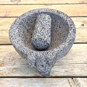 May include: A grey volcanic stone mortar and pestle set featuring a porous, textured surface. The circular mortar has a deep basin and a thick rim, while the cylindrical pestle rests inside the bowl, both displaying a natural speckled appearance.