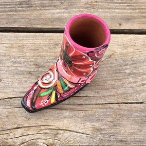 May include: A ceramic boot-shaped vessel featuring a vibrant hand-painted floral pattern in shades of red, pink, and black. The interior of the boot is a natural terracotta color, while the exterior rim is finished with a glossy bright pink glaze.