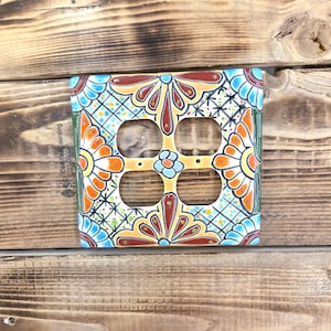 May include: A square ceramic light switch cover with a vibrant floral pattern. The cover showcases orange, blue, and yellow flowers with red and green details. It has four openings for electrical outlets.