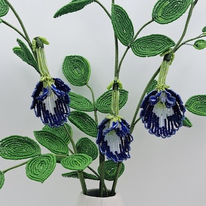 May include: A white vase holds beaded floral stems with green leaves and blue and white flowers. The beaded flowers and leaves are detailed and textured. The vase is a simple, modern design.