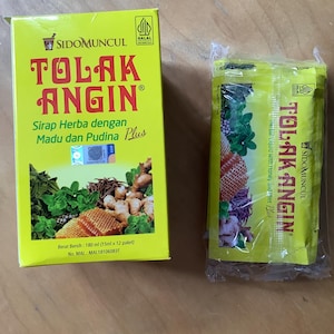 以下が含まれることがあります： 黄色い箱とTolak Anginハーブ液のパック。箱にはブランド名と製品情報が赤と緑のテキストで表示されています。パックは密封されており、同様のブランディングが表示されています。