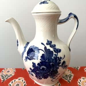 Puede incluir: Cafetera de porcelana blanca con diseño floral azul. La cafetera tiene tapa, boquilla curva y asa. El patrón floral es un diseño azul detallado sobre fondo blanco. La cafetera está sobre una superficie roja estampada.