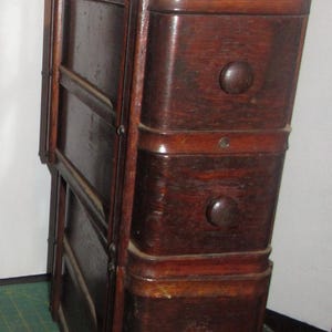 May include: A dark brown Art Deco style wooden cabinet with four drawers and round knobs. The cabinet is tall and narrow, with a hinged side. The text "2) Art Deco" is visible in the background.