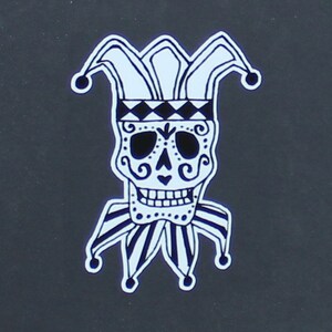 May include: A black and white illustration of a sugar skull wearing a jester's hat. The skull has intricate details and a whimsical expression.