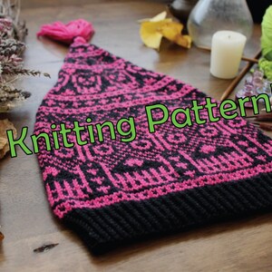 Può includere: Un cappello di lana rosa e nero con un motivo bianco e un pon pon rosa. Il cappello è appoggiato su una superficie di legno con il testo "Knitting Pattern!" sovrapposto all'immagine.