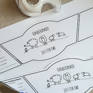 May include: Two white labels with black outlines and text that reads "Handcranked Just for Ewe" with a line drawing of sheep. The labels are on a beige surface with a pair of blue scissors and a roll of tape.