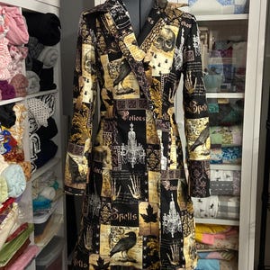 May include: A long, black and gold patterned coat with a V-neck collar. The coat features a repeating pattern of skulls, ravens, and the word "Spells". The coat is displayed on a mannequin.