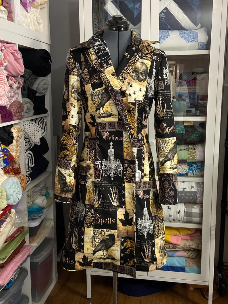 May include: A long, black and gold patterned coat with a V-neck collar. The coat features a repeating pattern of skulls, ravens, and the word "Spells". The coat is displayed on a mannequin.