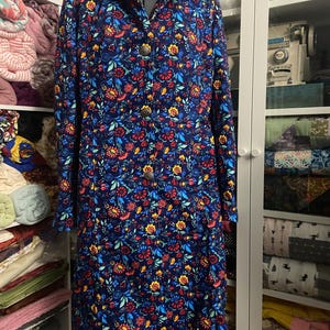 May include: A long-sleeved, knee-length dress with a vibrant floral pattern in red, yellow, and blue on a navy background. The dress features a collared neckline and is buttoned down the front with decorative buttons. The dress is displayed on a mannequin.