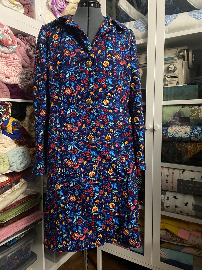 May include: A long-sleeved, knee-length dress with a vibrant floral pattern in red, yellow, and blue on a navy background. The dress features a collared neckline and is buttoned down the front with decorative buttons. The dress is displayed on a mannequin.