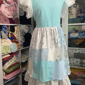 May include: A light blue dress with a patchwork skirt and white lace sleeves. The skirt features various blue and white patterned fabrics, including floral and geometric designs. The dress is displayed on a mannequin.