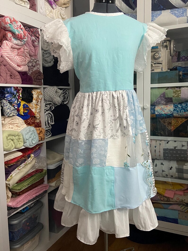 May include: A light blue dress with a patchwork skirt and white lace sleeves. The skirt features various blue and white patterned fabrics, including floral and geometric designs. The dress is displayed on a mannequin.