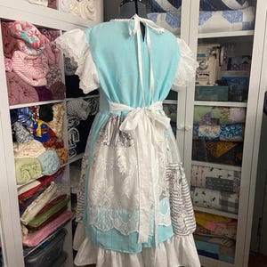 May include: A light blue dress with white lace sleeves and a white apron-style overlay. The dress features a white tie-back and a white waist tie. The skirt has a layered design with white ruffles. The dress is displayed on a dress form.