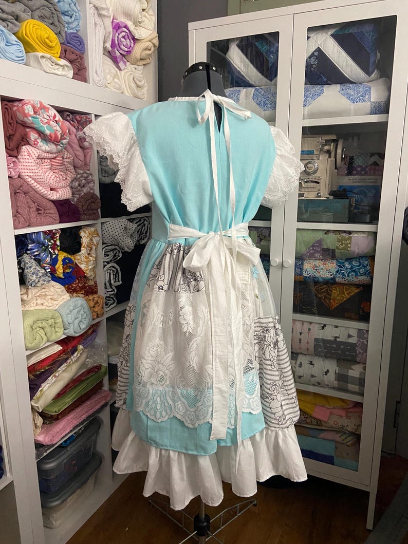 May include: A light blue dress with white lace sleeves and a white apron-style overlay. The dress features a white tie-back and a white waist tie. The skirt has a layered design with white ruffles. The dress is displayed on a dress form.