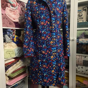 May include: A long-sleeved, button-down dress with a vibrant floral pattern in red, yellow, and blue on a navy background. The dress is displayed on a dress form, showcasing its tailored fit and design. The dress has a red trim on the cuffs.