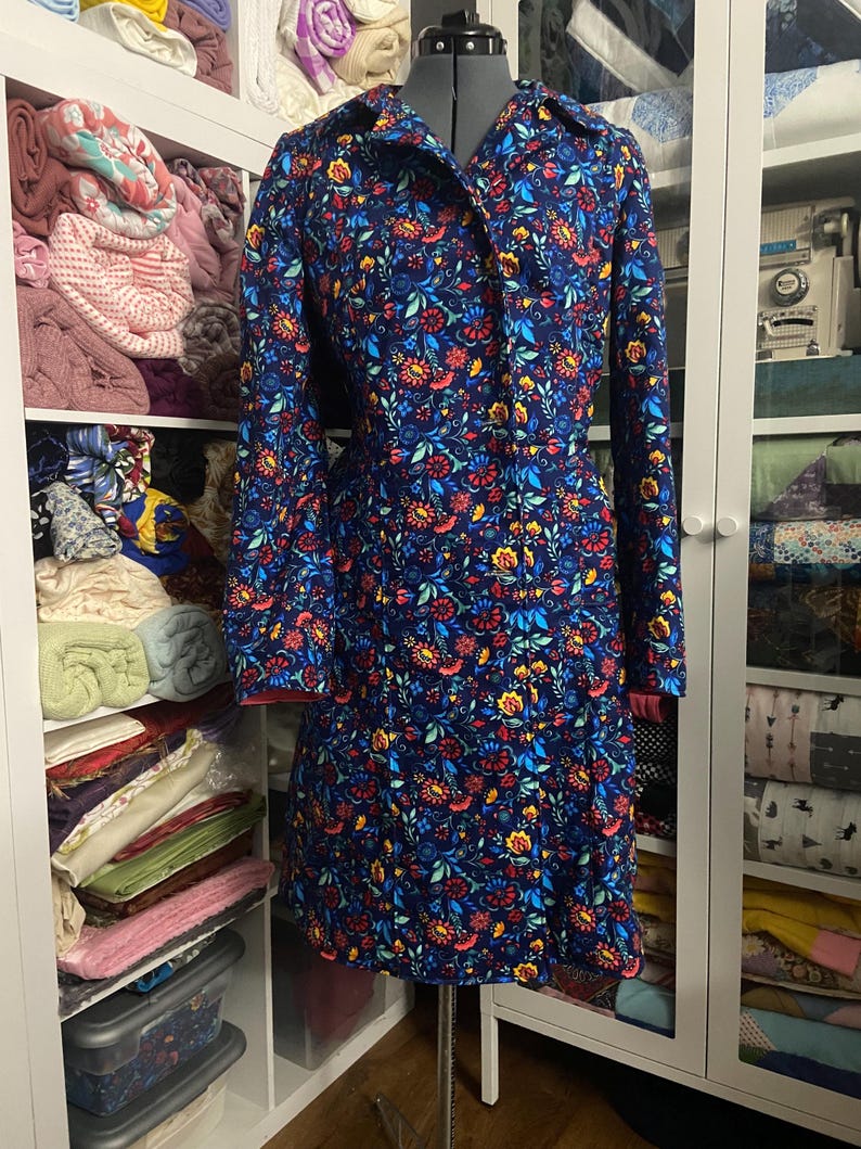 May include: A long-sleeved, button-down dress with a vibrant floral pattern in red, yellow, and blue on a navy background. The dress is displayed on a dress form, showcasing its tailored fit and design. The dress has a red trim on the cuffs.