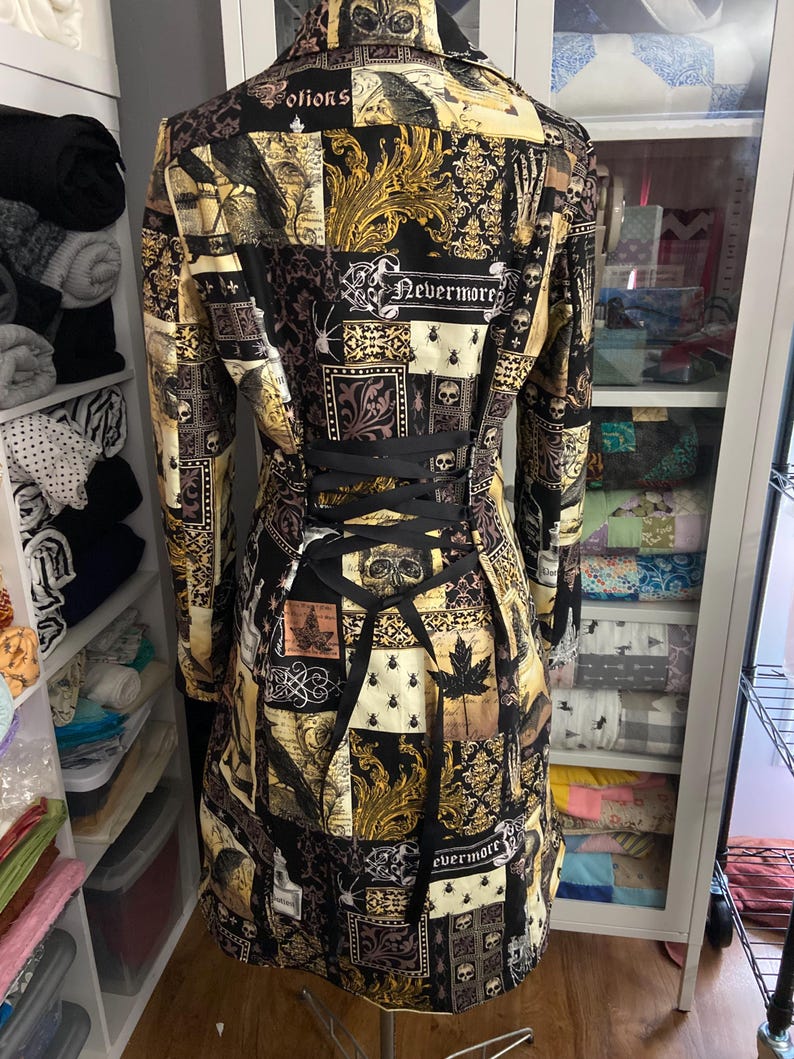 May include: A long-sleeved, patchwork-style coat with a black corset back. The coat features a collage of patterns in black, gold, and cream, including skulls, ornate designs, and the word "Nevermore".