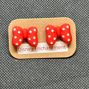 Pu&ograve; includere: Un paio di orecchini rossi a forma di fiocco con pois bianchi, presentati su un piccolo cartoncino rettangolare beige. Il cartoncino riporta la scritta "Disney Enchantment". Gli orecchini sono su uno sfondo grigio strutturato.