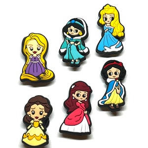 Puede incluir: Una colecci&oacute;n de coloridos dijes de zapatos de personajes de dibujos animados. Los dijes presentan a varias princesas con sus atuendos caracter&iacute;sticos, incluyendo a Rapunzel, Jasmine, Aurora, Blancanieves, Bella y otras. Cada dije tiene un borde negro.