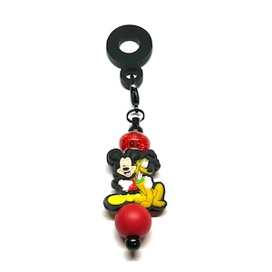 Puede incluir: Un dije negro con un diseño de Mickey Mouse y Pluto. El dije tiene una bola roja con purpurina, una bola roja y un anillo negro en la parte superior. Las figuras de Mickey Mouse y Pluto son en color.