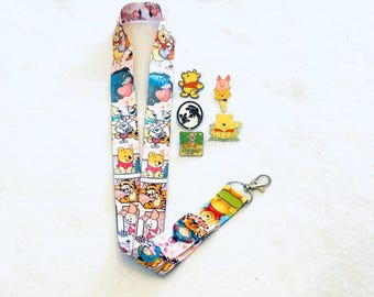 May include: A fabric lanyard featuring a repeating pattern of Winnie the Pooh characters including Tigger, Piglet, and Eeyore, paired with four metal enamel pins depicting various characters and the word Tigger in a green rectangular frame.