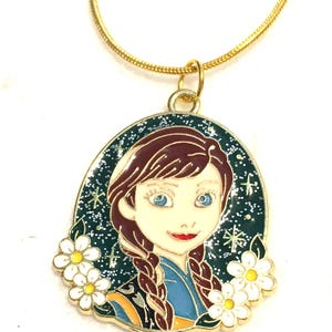 May include: A gold-toned necklace featuring a pendant with a cartoon image of a woman with brown braids, blue eyes, and a red lip. The pendant is surrounded by a dark green border with white flowers and a starry background.