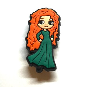 Puede incluir: Un dije de dibujos animados que representa a un personaje con cabello rizado naranja brillante y un vestido verde. El dije tiene una base negra y una expresi&oacute;n linda y amigable. Este accesorio es probablemente para zapatos u otros art&iacute;culos.