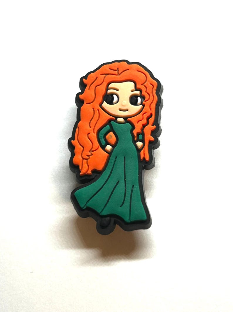 Puede incluir: Un dije de dibujos animados que representa a un personaje con cabello rizado naranja brillante y un vestido verde. El dije tiene una base negra y una expresi&oacute;n linda y amigable. Este accesorio es probablemente para zapatos u otros art&iacute;culos.