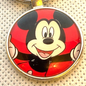 May include: A round keychain pendant featuring Mickey Mouse. The pendant has a red background with Mickey's face in the center. Mickey's ears are black, and his face is white with a smiling expression. The pendant is surrounded by a silver-colored metal frame.