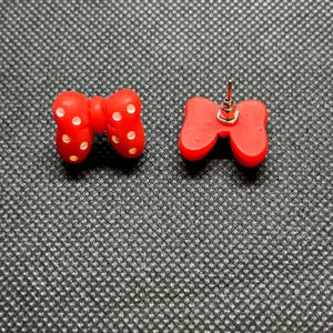 Pu&ograve; includere: Un paio di orecchini a bottone rossi a forma di fiocco. Ogni orecchino presenta un design a fiocco con pois bianchi. Gli orecchini sono realizzati in materiale lucido e sono su sfondo grigio strutturato.