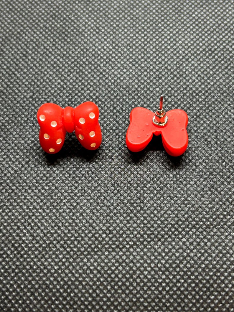 Pu&ograve; includere: Un paio di orecchini a bottone rossi a forma di fiocco. Ogni orecchino presenta un design a fiocco con pois bianchi. Gli orecchini sono realizzati in materiale lucido e sono su sfondo grigio strutturato.