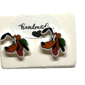 May include: A pair of Pluto character earrings. Each earring features a cartoon image of Pluto with a brown head, black ears, and a red tongue. The earrings are on a white card with the word "Handmade" written on it.