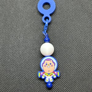 May include: A blue and white keychain featuring a cartoon astronaut character. The keychain has a blue loop, a small chain, a white bead, and a cartoon astronaut figure with a blue and purple helmet. The astronaut has a yellow and green outfit.