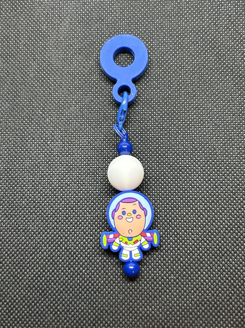 May include: A blue and white keychain featuring a cartoon astronaut character. The keychain has a blue loop, a small chain, a white bead, and a cartoon astronaut figure with a blue and purple helmet. The astronaut has a yellow and green outfit.
