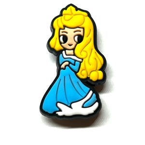 Puede incluir: Un charm de princesa de dibujos animados con un vestido azul con ribetes blancos y un contorno negro. La princesa tiene el pelo largo y amarillo. El charm probablemente est&aacute; hecho de goma o un material similar.