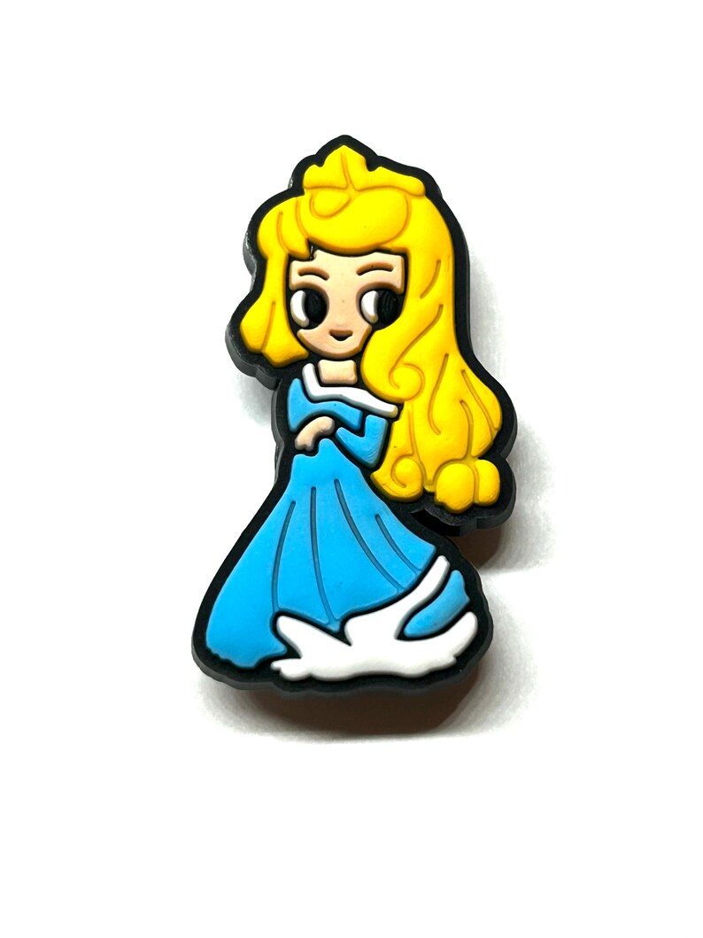 Puede incluir: Un charm de princesa de dibujos animados con un vestido azul con ribetes blancos y un contorno negro. La princesa tiene el pelo largo y amarillo. El charm probablemente est&aacute; hecho de goma o un material similar.