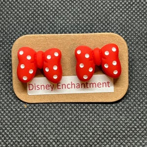 Pu&ograve; includere: Un paio di orecchini rossi a forma di fiocco con pois bianchi, presentati su una scheda rettangolare marrone. La scheda riporta la scritta "Disney Enchantment". Gli orecchini sono su uno sfondo grigio.