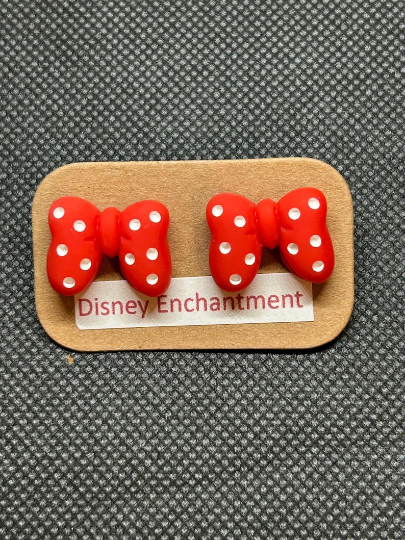 Pu&ograve; includere: Un paio di orecchini rossi a forma di fiocco con pois bianchi, presentati su una scheda rettangolare marrone. La scheda riporta la scritta "Disney Enchantment". Gli orecchini sono su uno sfondo grigio.