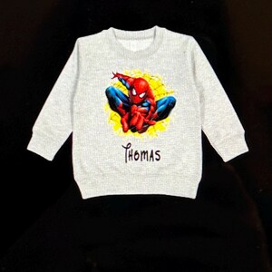 K&ouml;nnte beinhalten: Hellgraues Sweatshirt mit einem farbenfrohen Spider-Man-Motiv. Der Superheld ist in seinem klassischen rot-blauen Anzug vor einem gelben, spinnennetzartigen Hintergrund dargestellt. Der Name "Thomas" ist darunter aufgedruckt.