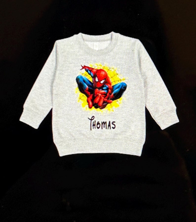 K&ouml;nnte beinhalten: Hellgraues Sweatshirt mit einem farbenfrohen Spider-Man-Motiv. Der Superheld ist in seinem klassischen rot-blauen Anzug vor einem gelben, spinnennetzartigen Hintergrund dargestellt. Der Name "Thomas" ist darunter aufgedruckt.