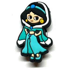 Puede incluir: Un amuleto colorido con un personaje de dibujos animados con un vestido turquesa y cabello negro. El amuleto tiene un borde blanco y aretes dorados. Una gema azul est&aacute; en la cabeza del personaje. El amuleto es un accesorio para zapatos.