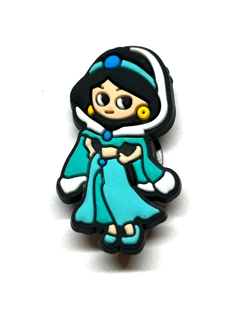 Puede incluir: Un amuleto colorido con un personaje de dibujos animados con un vestido turquesa y cabello negro. El amuleto tiene un borde blanco y aretes dorados. Una gema azul est&aacute; en la cabeza del personaje. El amuleto es un accesorio para zapatos.