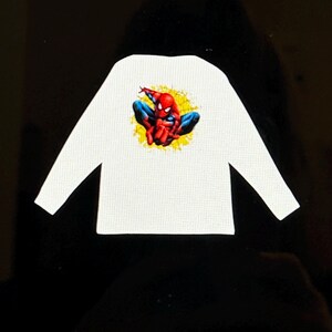 K&ouml;nnte beinhalten: Wei&szlig;es Langarmshirt mit einer farbenfrohen Grafik von Spider-Man in einer hockenden Pose. Der Superheld ist in seinem klassischen rot-blauen Anzug vor einem gelben Hintergrund dargestellt. Das Shirt hat ein strukturiertes Muster.