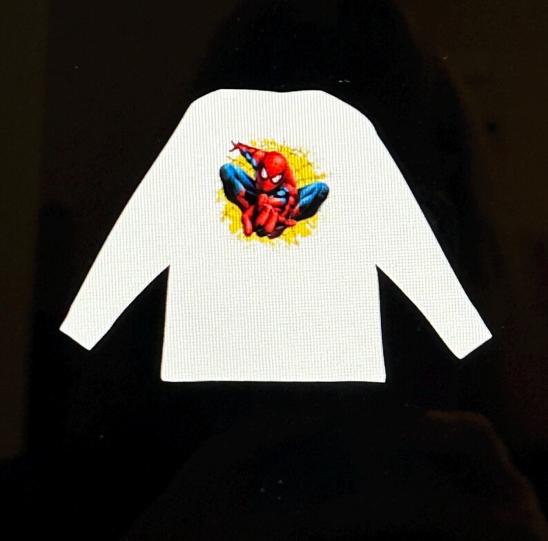 K&ouml;nnte beinhalten: Wei&szlig;es Langarmshirt mit einer farbenfrohen Grafik von Spider-Man in einer hockenden Pose. Der Superheld ist in seinem klassischen rot-blauen Anzug vor einem gelben Hintergrund dargestellt. Das Shirt hat ein strukturiertes Muster.