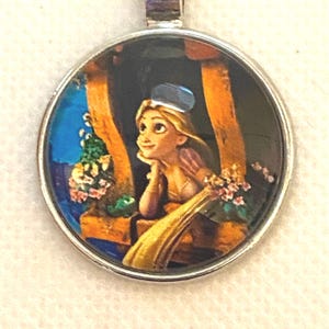 May include: A circular silver pendant with a cartoon illustration of a young woman with long blonde hair looking out of a window. The image includes flowers, a blue sky, and wooden window frames. The pendant is attached to a silver chain.
