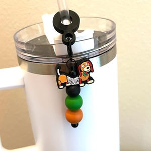 May include: A white tumbler with a black straw holder and a decorative charm. The charm features a cartoon dog with orange, black, and green beads. The tumbler has a clear lid and a silver band.