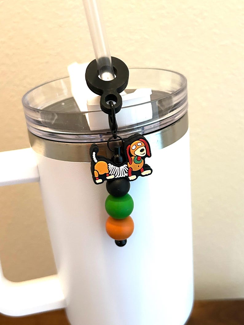May include: A white tumbler with a black straw holder and a decorative charm. The charm features a cartoon dog with orange, black, and green beads. The tumbler has a clear lid and a silver band.