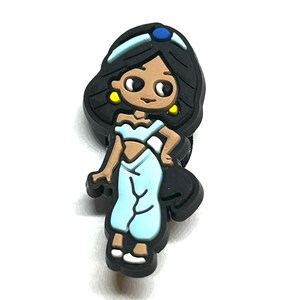 Puede incluir: Un dije de estilo caricatura que presenta un personaje con cabello negro, piel morena y ropa azul claro. El dije tiene una diadema azul con una gema azul, aretes amarillos y zapatos blancos. El personaje est&aacute; de pie.